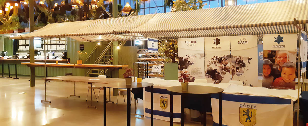 A large white tent is set up in a room with a dining table underneath it. The tent is open and has a blue and yellow sign on it. There are several chairs placed around the room, and a potted plant is located near the center of the room. AI generated content