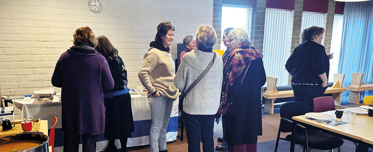 A group of people are standing around a table, engaged in conversation. There are several chairs placed around the table, and a clock on the wall. The people are of various ages and genders, and some are wearing ties. The atmosphere appears to be social and friendly. AI generated content