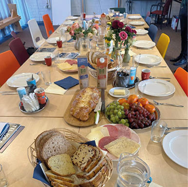 A table is set with a variety of food and drinks, including bread, grapes, and juice. There are also plates, cups, and bottles on the table. A person is standing near the table, and a chair is visible in the background. AI generated content