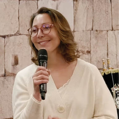 A person with shoulder-length hair wearing glasses and a white cardigan is holding a microphone and standing in front of a stone wall with a decorative object partially visible behind them. Description generated by AI