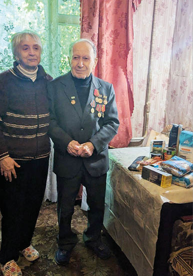Two seniors stand side by side in a room with floral curtains, one wearing a suit adorned with medals and the other in a dark sweater, next to a table covered with various packaged food items. Description generated by AI