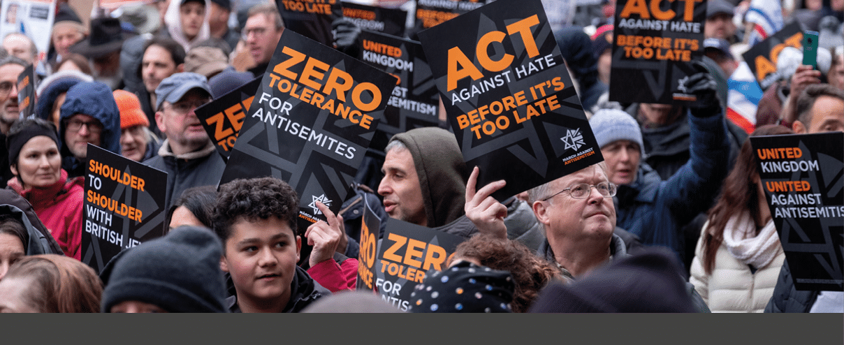 A crowd of people outdoors hold signs with messages against hate and antisemitism, including phrases like “Zero tolerance for antisemites" and "Act against hate before it's too late." Description generated by AI