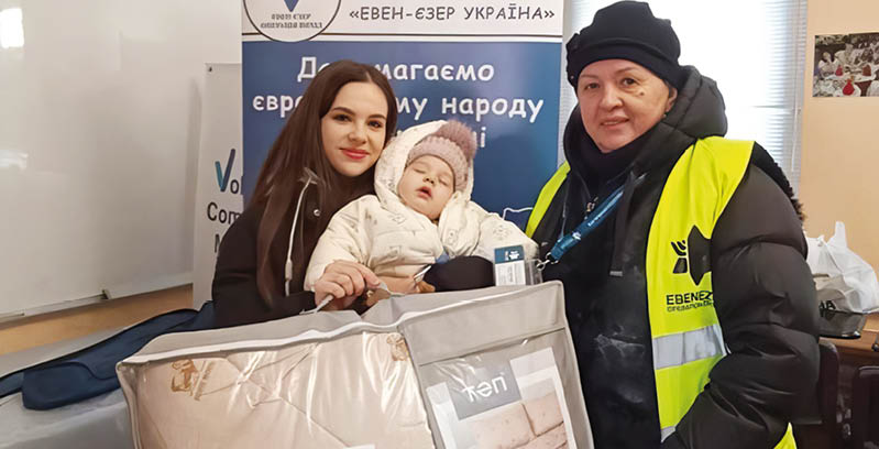 Two adults and a child are indoors with one adult wearing a yellow safety vest and the child bundled in warm clothing while sitting in a large padded carrier. Description generated by AI