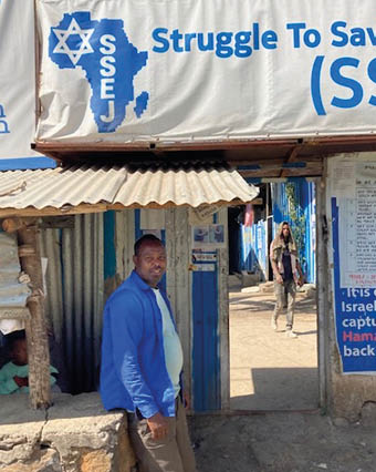 A person in a blue jacket stands near a small structure under a sign that reads “Struggle To Save" with an outline of Africa and text including "SSEJ," while another person walks in the background along a sunlit street. Description generated by AI