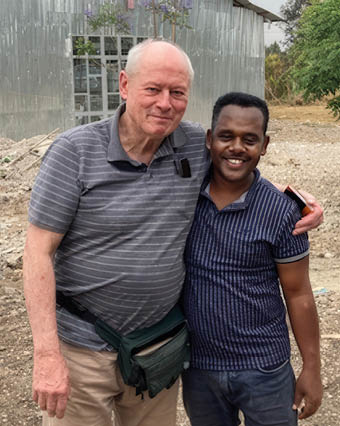 Two adults stand closely together outdoors with one arm around the other, smiling in front of a metal building and a dirt ground. Description generated by AI
