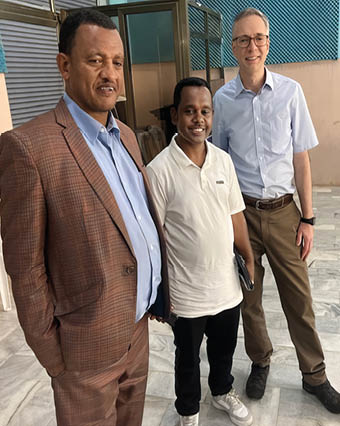 Three adults stand indoors on a marble floor, with one wearing a brown checked suit, another in a white polo shirt holding a tablet, and the third in a light blue shirt and brown pants smiling. Description generated by AI