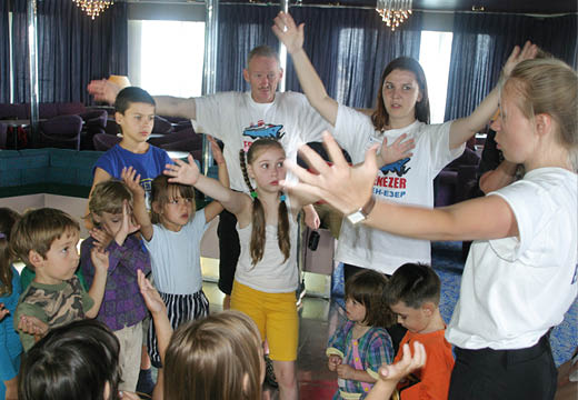 A group of children and adults stand in a circle indoors with their arms raised, appearing to participate in an interactive activity or game. Description generated by AI