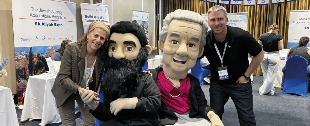 Two adults pose smiling next to two large seated puppets with exaggerated facial features at an indoor event with informational booths in the background. Description generated by AI