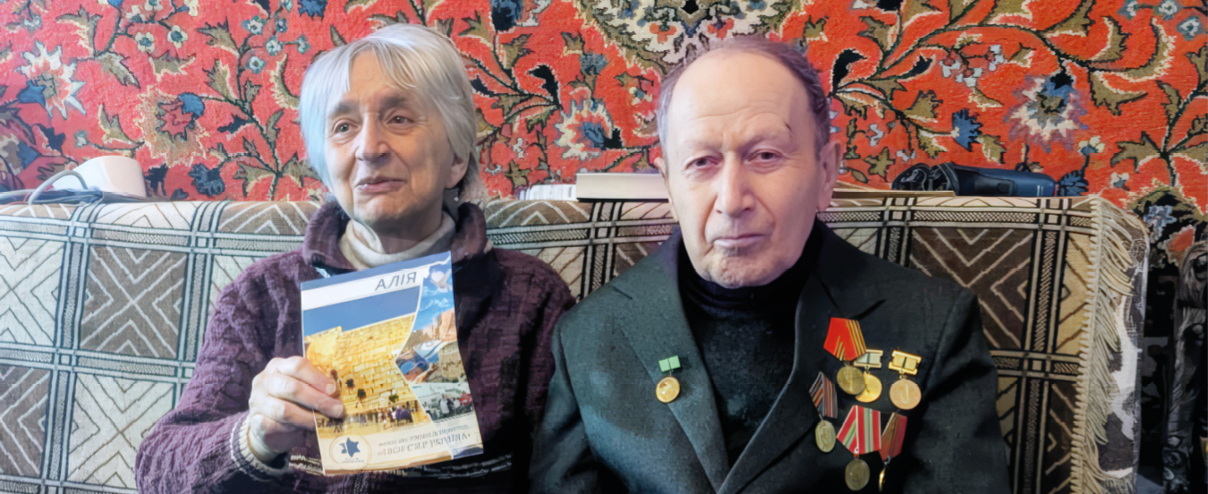 Two seniors sit side by side on a patterned couch with a colorful carpet hanging behind them, one holding a booklet and the other wearing a jacket adorned with medals. Description generated by AI