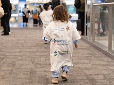 A child with curly hair wearing a long white shirt with text and logos walks down a carpeted hallway with other children and adults in the background. Description generated by AI