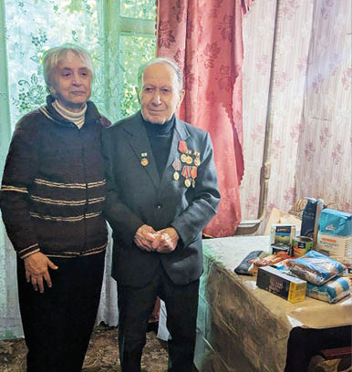 Two seniors stand side by side in a room with floral curtains, one wearing a suit adorned with medals and the other in a dark sweater, next to a table covered with various packaged food items. Description generated by AI