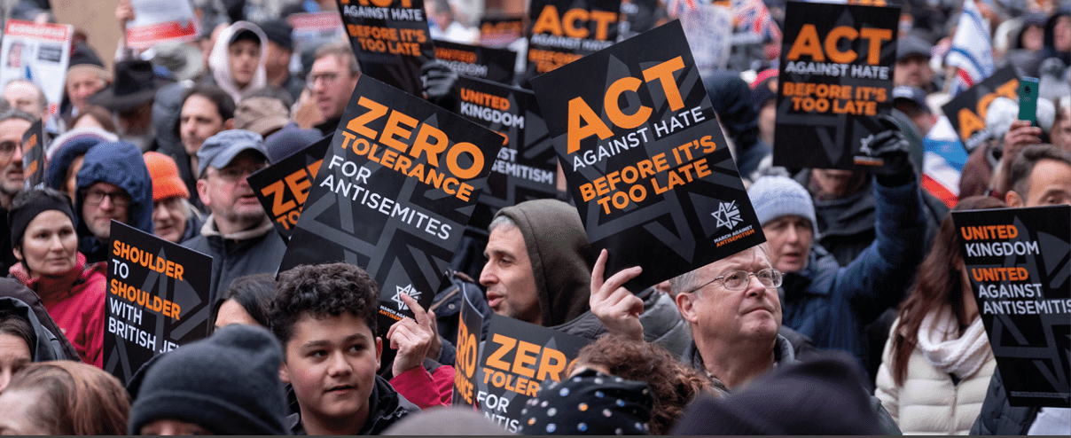 A crowd of people outdoors hold signs with messages against hate and antisemitism, including phrases like “Zero tolerance for antisemites" and "Act against hate before it's too late." Description generated by AI
