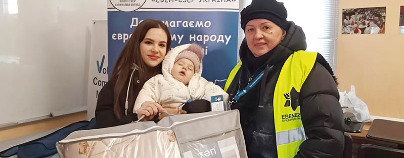 Two adults and a child are indoors with one adult wearing a yellow safety vest and the child bundled in warm clothing while sitting in a large padded carrier. Description generated by AI