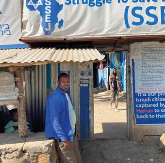 A person in a blue jacket stands near a small structure under a sign that reads “Struggle To Save" with an outline of Africa and text including "SSEJ," while another person walks in the background along a sunlit street. Description generated by AI