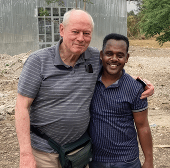 Two adults stand closely together outdoors with one arm around the other, smiling in front of a metal building and a dirt ground. Description generated by AI