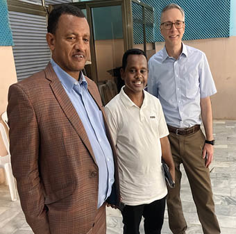 Three adults stand indoors on a marble floor, with one wearing a brown checked suit, another in a white polo shirt holding a tablet, and the third in a light blue shirt and brown pants smiling. Description generated by AI