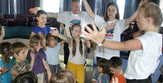 A group of children and adults stand in a circle indoors with their arms raised, appearing to participate in an interactive activity or game. Description generated by AI