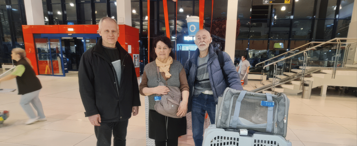 Three adults stand indoors near a pet carrier and a soft-sided pet travel bag in a brightly lit public space with large windows and stairs in the background. Description generated by AI