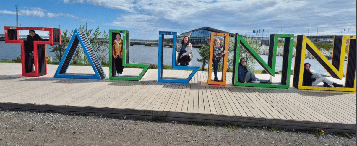 Large colorful letters spelling “TALLINN" are arranged outdoors on a wooden platform near water, with several people sitting or standing inside the hollow letters under a partly cloudy sky. Description generated by AI