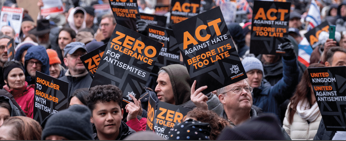A crowd of people outdoors hold signs with messages against hate and antisemitism, including phrases like “Zero tolerance for antisemites" and "Act against hate before it's too late." Description generated by AI