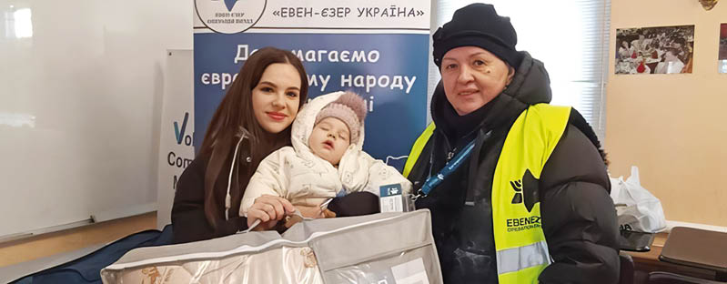 Two adults and a child are indoors with one adult wearing a yellow safety vest and the child bundled in warm clothing while sitting in a large padded carrier. Description generated by AI