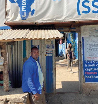 A person in a blue jacket stands near a small structure under a sign that reads “Struggle To Save" with an outline of Africa and text including "SSEJ," while another person walks in the background along a sunlit street. Description generated by AI