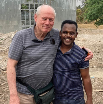 Two adults stand closely together outdoors with one arm around the other, smiling in front of a metal building and a dirt ground. Description generated by AI
