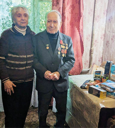 Two seniors stand side by side in a room with floral curtains, one wearing a suit adorned with medals and the other in a dark sweater, next to a table covered with various packaged food items. Description generated by AI