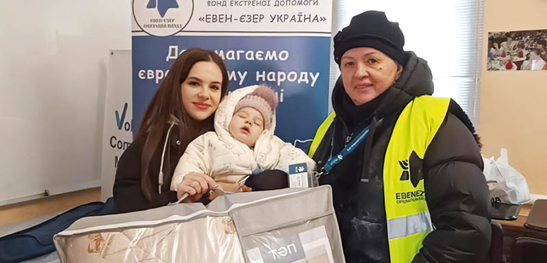 Two adults and a child are indoors with one adult wearing a yellow safety vest and the child bundled in warm clothing while sitting in a large padded carrier. Description generated by AI