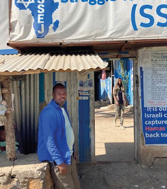 A person in a blue jacket stands near a small structure under a sign that reads “Struggle To Save" with an outline of Africa and text including "SSEJ," while another person walks in the background along a sunlit street. Description generated by AI