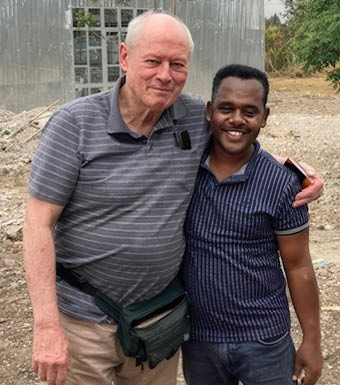 Two adults stand closely together outdoors with one arm around the other, smiling in front of a metal building and a dirt ground. Description generated by AI