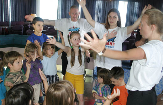 A group of children and adults stand in a circle indoors with their arms raised, appearing to participate in an interactive activity or game. Description generated by AI