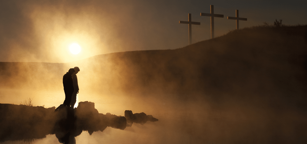 Dramatic religious photo illustration of Easter Sunday Morning reflecting a prayerful moment of silence with a silhoutted person bowing his head, a warm sunrise rises over a foggy lake, and three crosses appear as a vision on a hill, reflected in the water as well 