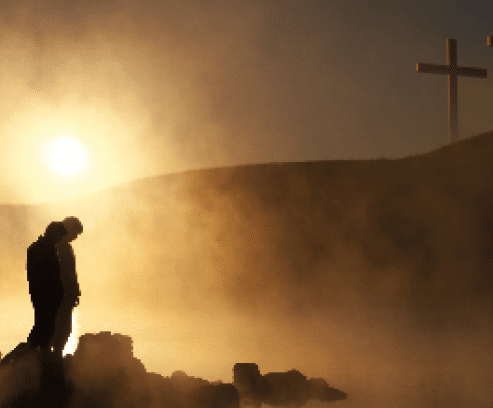 Dramatic religious photo illustration of Easter Sunday Morning reflecting a prayerful moment of silence with a silhoutted person bowing his head, a warm sunrise rises over a foggy lake, and three crosses appear as a vision on a hill, reflected in the water as well 