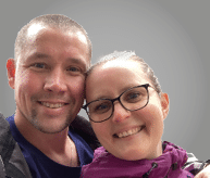 Two adults wearing casual jackets are standing close together against a plain gray background. Description generated by AI